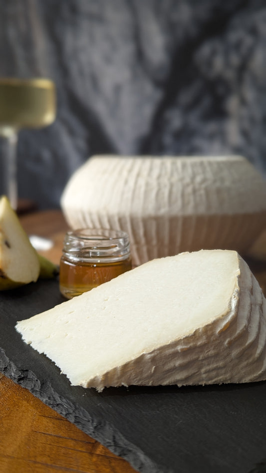 A wedge of creamy off-white goats cheese on a dark slate board, with a small glass jar of honey and a pear slice in a softly blurred kitchen or bar setting.