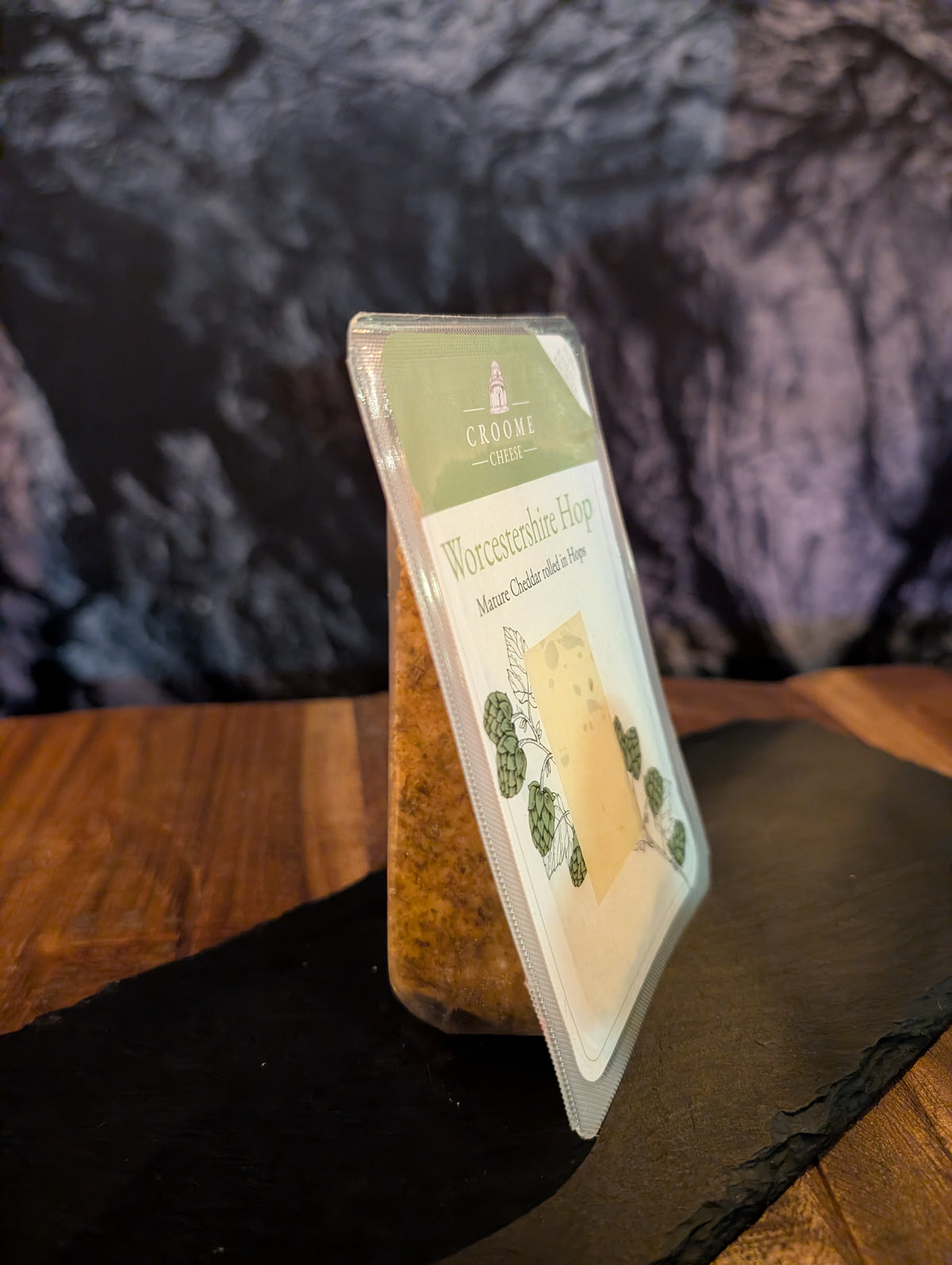 A package of Croome Worcestershire hop seasoning, shown standing upright on a dark slate board over a wooden table with a dark, textured background