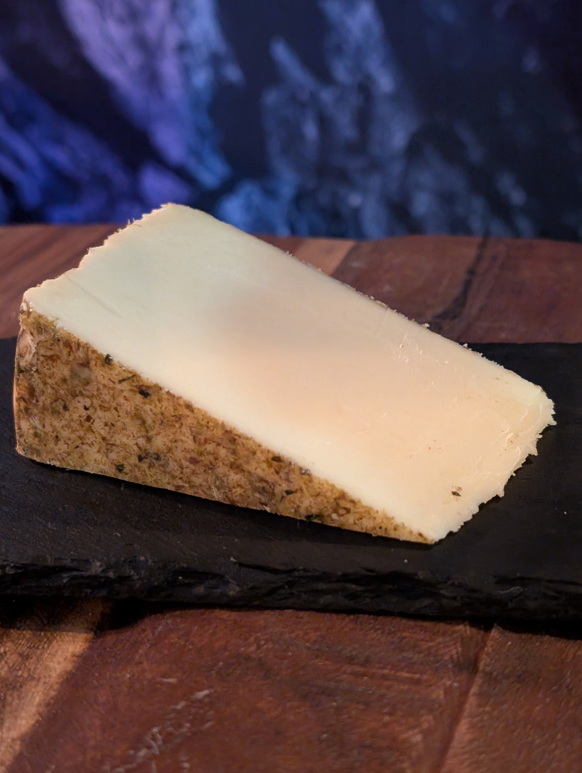 A wedge of semi-hard cheese with a pale creamy interior and a textured brown rind, displayed on a dark slate board over a wooden surface.