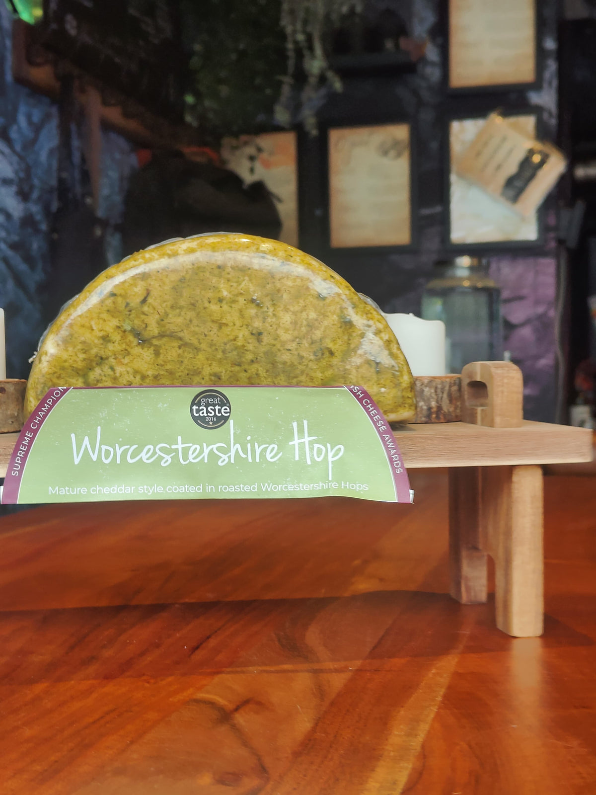A packaged cheese wedge labeled 'Worcestershire Hop' displayed on a small wooden stand on a wooden table, with a blurred indoor background.