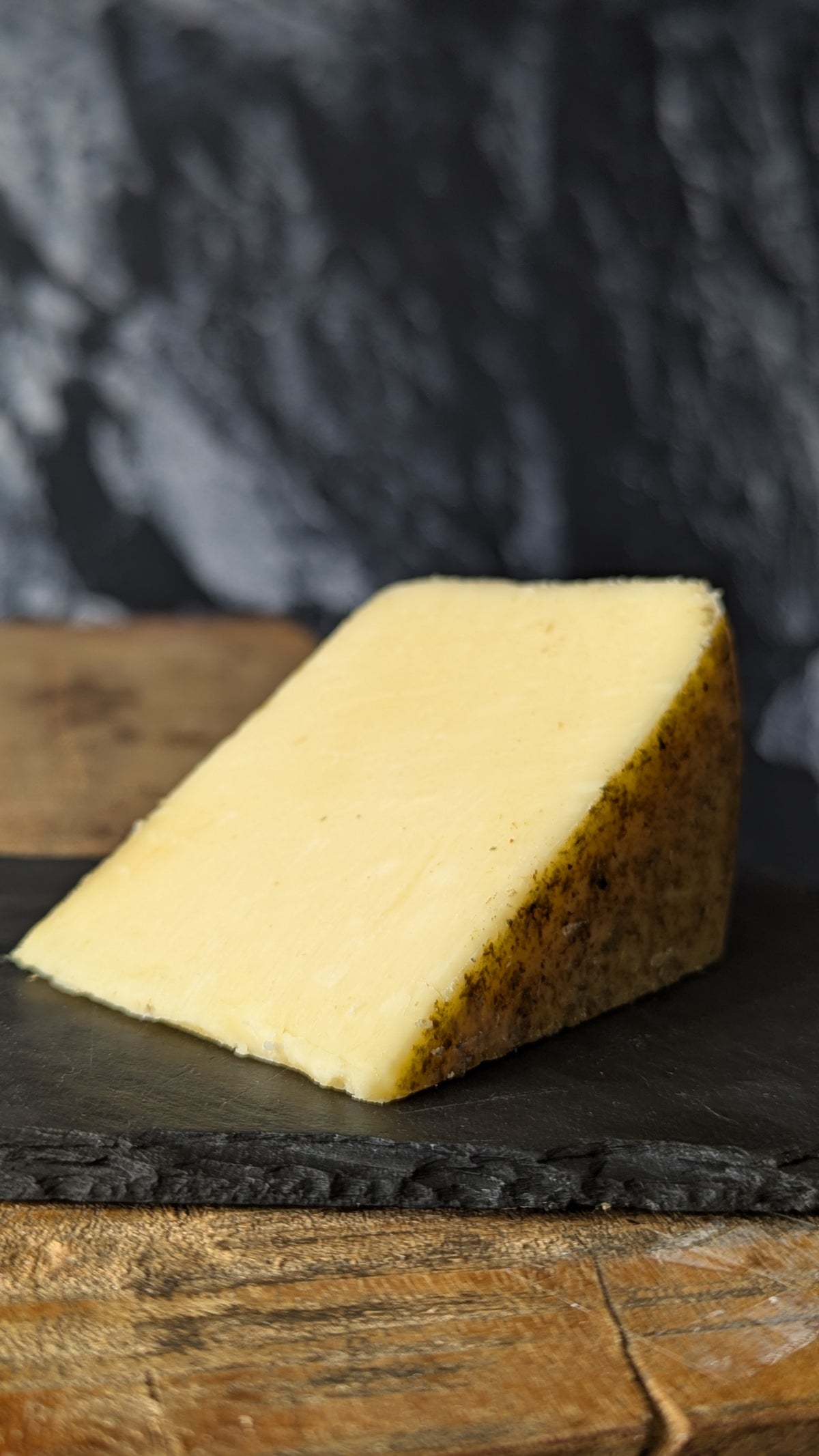 A wedge of pale yellow cheese with a textured golden-brown rind, resting on a dark slate board over a rustic wooden surface against a dark blurred background.