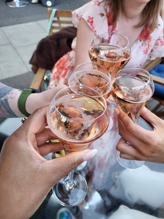 Four hands holding champagne flutes filled with rose gold wine, clinking together in a toast at the local cheese cave. A woman in a pink floral dress is seated in the background.