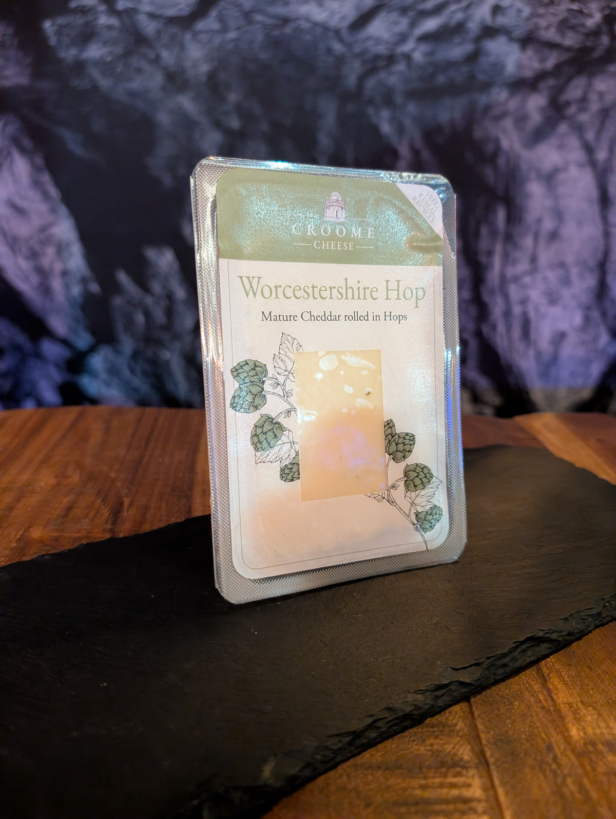 A packaged cheese puck labeled 'Worcestershire Hop' by Croome Cheese, standing upright on a dark slate board over a wooden surface with a dark textured backdrop.
